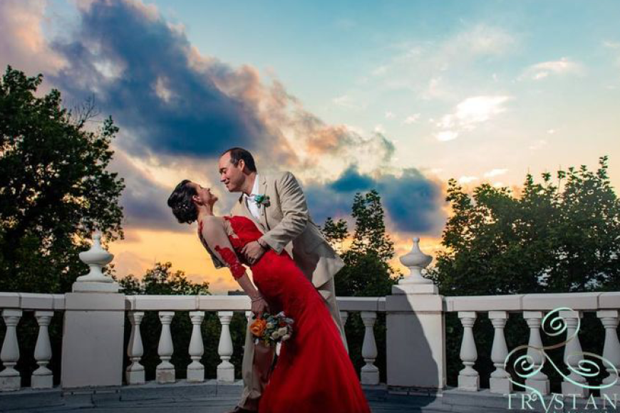 A newlywed couple in front of Grant Humphreys mansion, the bride is being dipped by the groom at sunset. She is in a striking red dress.