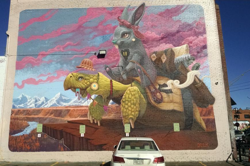 Colorful brick mural showing a fantasy version of colorado. A black anthropomorphic bunny rides a turtle with a hat and saddle on.