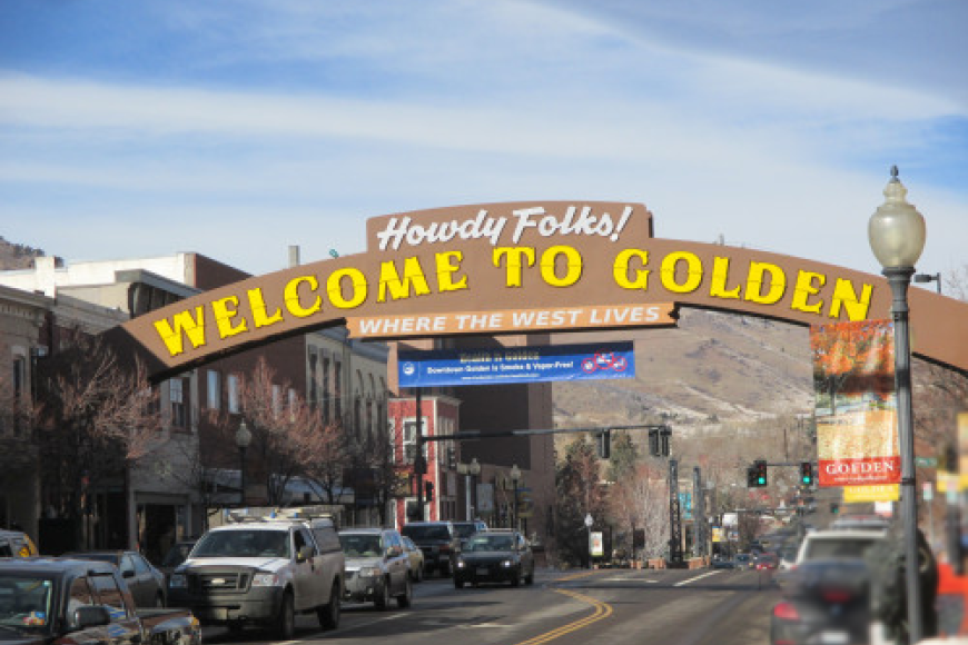 Main street golden lined with business and cars. The Large orange and brown sign reads "Howdy Folks! Welcome to Golden. Where the west lives"