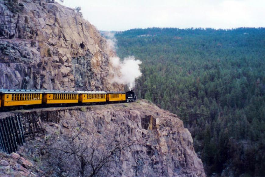 A yellow and green train moving along a narrow cliff edge in Colorado.