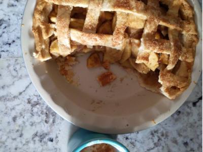 Photo looking directly down at a homemade apple pie, sitting atop a gray and white granite countertop. The pie has been baked in a white stoneware pie plate, and about one-third of the pie is already gone. The pie is apple, with a golden lattice top that is sprinkled with sugar. Next to the pie plate, there is a turquoise coffee cup that had latte in it, although nearly all of the coffee has been enjoyed and just the foam, sprinkled with cinnamon, remains.
