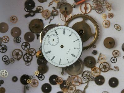 Photo of a white clock face with thin black roman numerals. The clock is in pieces, and all of the small cogs that make the clock function are scattered around in the area around the clock face.  The hands on the clock face are missing.