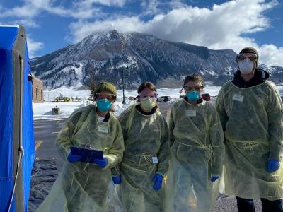 Photo of a Covid-19 testing center in Crested Butte. There are 4 medical professionals dressed in gowns, safety glasses, and masks, and blue nitrile gloves, standing in a parking lot. There is snow visible on the ground and the mountain behind them.