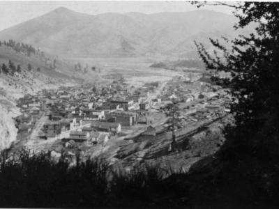 Photo of the town of Creede in the year 1918. The image is taken from atop a hill overlooking the town, so trees and brush frame the view of the town in the distance. Two main streets through town are evident, and low buildings of small to mid-sized are arranged through the town. No vehicles appear to be seen, and the town is too far away to see people walking around..