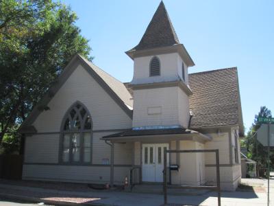 Photo of the front view of People's Methodist Episcopal Church. The building is wooden with shingled sides that are painted a tan color. The trim work is a medium brown color. There are simple wooden double doors in the front which are painted white and have twin rectangular windows. There is one large arched window to the left of the front doors,  and a small steeple directly above the  front doors. The church sits on the corner of Saint Vrain and another street, right next to the sidewalks.