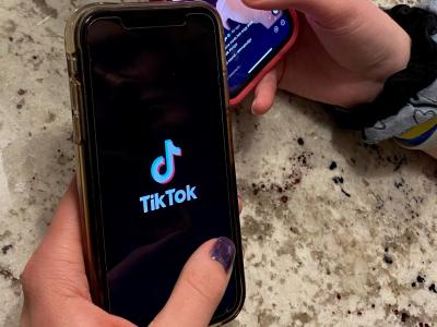 Photo of two people holding cell phones that are open to the TikTok app. One phone has the TikTok logo, and the other phone shows a photo of a man and his cat--the cat is wearing a hat and meowing.