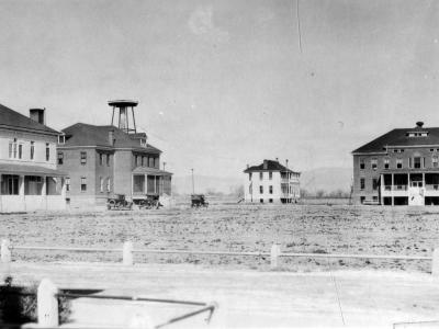 Photo of four buildings sitting on a barren piece of land. in the foreground, part of a dirt road can be seen. There are 3 vehicles parked in front of two of the buildings on the left side of the image. In the far distance, the hazy silouette of mountains can be seen. There are no people immediately visible in this historic photograph.