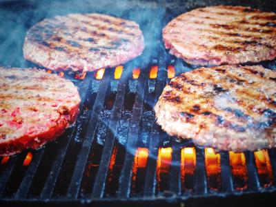 Photo closeup of four beef hamburgers cooking on a grill.  The first side of the burgers has been cooked, because the tops of each burger patty have "grill marks" lining them. The orange grill flames are visible beneath the black cast iron grates of the grill and a few wisps of smoke are rising from the cooking surface.