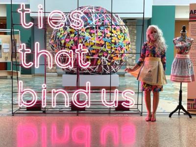 Photo of EllenAnne Geisel, standing next to a large neon sign with pink letters, that reads "ties that bind us." EllynAnne is wearing a bright floral pattern dress and a cheery yellow apron, and Covid-19 face mask. She is standing next to a mannequin that is also dressed in an apron, and all are in the atrium of the History Colorado Center in Denver.