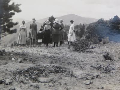 Photo of a group of women standing at the edge of a hill overlooking the mountains and valleys. Two of the women are holding the hand of a small Black boy, one on either side of him. He, too, is looking at the scenery. The women in this historic photo are all wearing dresses and most of them wear wide-brimmed hats. The young boy is wearing white shorts and a dark-colored shirt with a white collar.