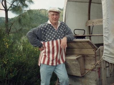 Photo of a man standing on the front hitch of a wooden covered wagon that is outdoors, surrounded by green shrubs and trees. He is wearing brightly colored shoes, blue jeans, and a white flat cap. His casual, rugby-style shirt represents the American flag: the top half of the shirt is blue with white stars, while the bottom half is red and white vertical stripes, and the collar is white. He is leaning his left elbow on the wagon and he is looking at the camera.
