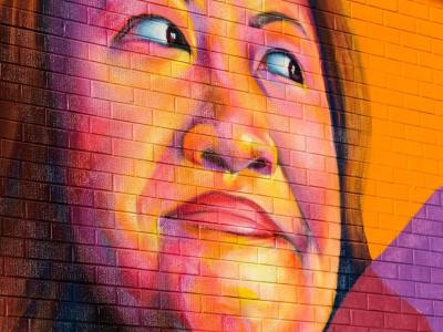 Photo of a mural painted on an exterior brick wall, of refugee and advocate Nga Vương-Sandoval. The image is a close up of her face, smiling. It is brightly colored in oranges, pinks, and reds. The mural was painted by artist/muralist Thomas Evans (I Am Detour).