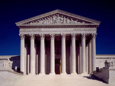 Photo of the west pediment of the Supreme Court Building in Washington DC.  The Image shows the monumental building of white marble, with its columns and massive staircase leading up to the door. Seated statues are seen toward the top of either side of the staircase. Above the columns, the pediment features a sculptural group of nine figures along with the words, "Equal Justice Under Law."