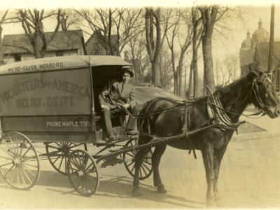 Photo of a horse-drawn car. The side of the car has the words "Volunteers of America - Relief Dept." painted on the side, along with "Phone Maple 790." The driver is a young man dressed in a suit and hat. He is sitting in the car,holding the reins and posing for this photo.