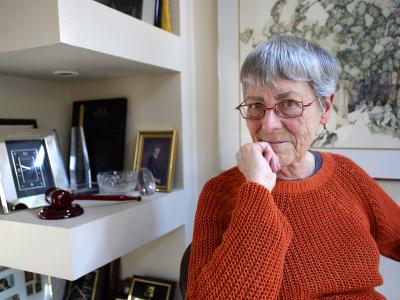 Photo of Jean Dubofsky, sitting in her office. The walls and shelves behind her are white, contrasting her orange sweater and red-framed eyeglasses. There are gold and silver-framed photos on the shelves, and a painting on the wall behind her. She is resting her chin on her closed right hand, and she is looking straight ahead at the camera.