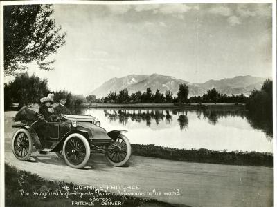 A man and a woman in turn-of-the-century dress ride in an early open-top car. The car is driving along a gravel path next to a small body of water. Mountains are on the horizon. A caption reads "The 100-mile Fritchle: The recognized highest-grade electric automobile in the world. Address: Fritchle, Denver."