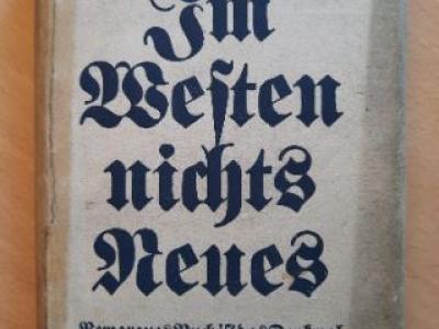 Photograph of a book with a worn white cover. The words on the cover are all in German, and in the center in large black letters the cover of the book reads, "Im Westen nichts Neues" which translates to "All Quiet on the Western Front."  Red block letters across the top of the book spell out the author's name, Erich Maria Remarque. 