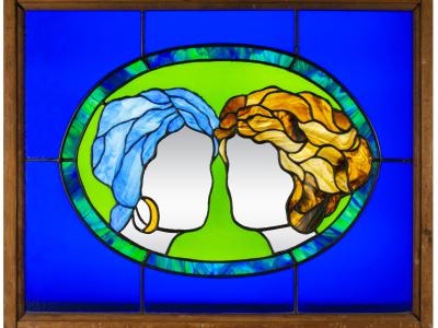 A stained glass window, depicting two women's heads facing each other. One has blue hair, the other has blonde hair. They are faceless.