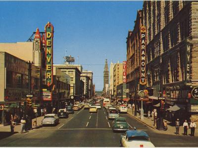 Image of a picture postcard of Downtown Denver around 1950-1960. The photograph has been taken from an intersection along Sixteenth Street. Signs on buildings read "Paramount (Theatre)" "Denver" "Skagg's" and "Swift's Ice Cream," among others. Cars are travelling in one direction along Sixteenth, toward the D&F Tower in the distance.