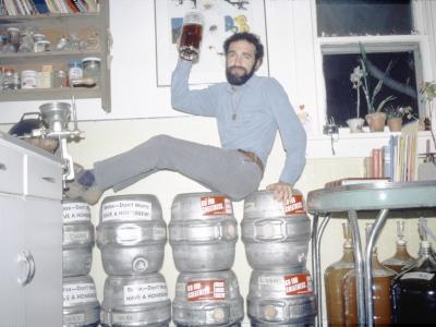Charlie Papazian posing on a group of kegs. He holds a mug of beer, and jugs of brewing beer are visible underneath a nearby table.