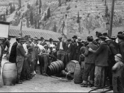 A group of men in period 1920s clothing stand around a wooden barrel of alcohol while it is poured into the gutter.