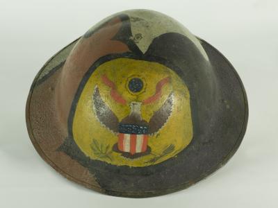 Photo of a Doughboy helmet from World War One. It is a round metal helmet painted with green, brown and black camouflage pattern on the outer surface. A heraldic eagle painted on a yellow background is hand painted on the forehead.