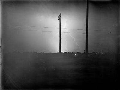 Photo of a large lightening bolt coming out of the cloud in the middle of the sky, and several fingers of lightening contact the ground. There are two very tall power line poles in the foreground. The bright lightening illuminates the darkness in this very dark photograph.