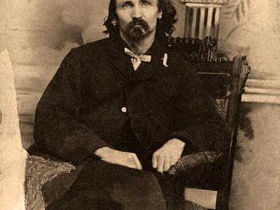 Alfred Packer, seated in a chair. He wears 19th century clothes and a scraggly goatee.