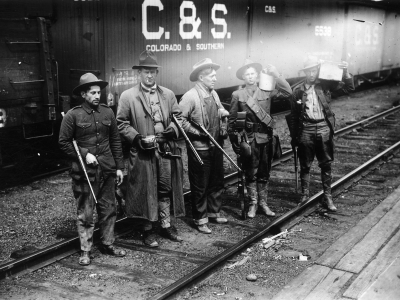 Four men standing on rail road tracks in front of a box car labelled C&S. The men are wearing hats and holding rifles.