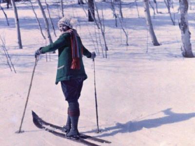A woman stands on two skis in a snowy landscape. She is wearing a jacket, scarf, and hat, and looks over her shoulder at the camera.