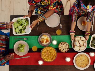 Photo looking down upon a long dining table made of dark brown wood and decorated with a long table runner down the center consisting of wide green and red stripes divided by a thin black stripe of fabric. Three people in colorful attire sit around the table, passing dishes of food and serving some onto plates. Dishes of greens, cornbread, green beans, sliced bread, and other foods are being shared.