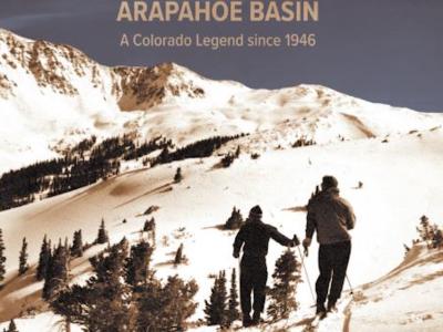 Image of the front cover a book titled "Arapahoe Basin: A Colorado Legend Since 1946." The authors, whose names appear at the bottom in small font, are Cathleen Norman and Alan J. Henceroth. The cover is a photograph of two skiers traversing a snow-covered hillside while looking at the snowy mountain in the distance. The sky is a brilliant blue, with no clouds in sight.