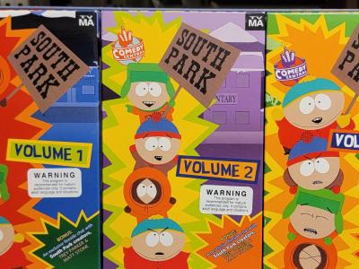 A photograph taken with a cell phone camera, of three VHS tapes in their cardboard covers, lined up on white metal shelving so that the full cover of each tape is visible. The tapes are recordings of the "South Park" animated television series, Volumes 1, 2, and 3. Each cover prominently displays the "TV-MA" warning in the upper right corner, as well as a warning box below the volume number, indicating that these programs are for mature, audiences only and contain adult language and situations.