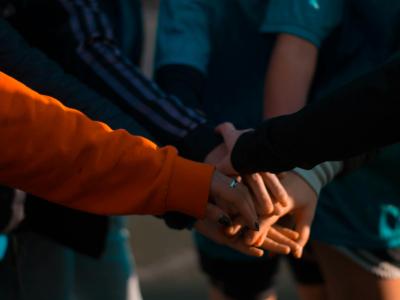 Photograph of a group of people who are stacking up their hands as if in a team going to play a game, or a group of people getting ready to undertake a team task. The sun shines on their hands, and several of the people wear long-sleeved colorful clothing.