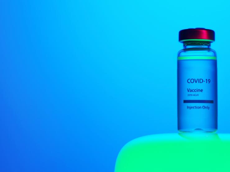Photo of one vial of Covid-19 Vaccine. The Vial has a red metal top and a label that says "COVID-19 Vaccine, 2019 n-CoV, Injection Only." The vial sits atop a green table, and the background is a luminescent blue.