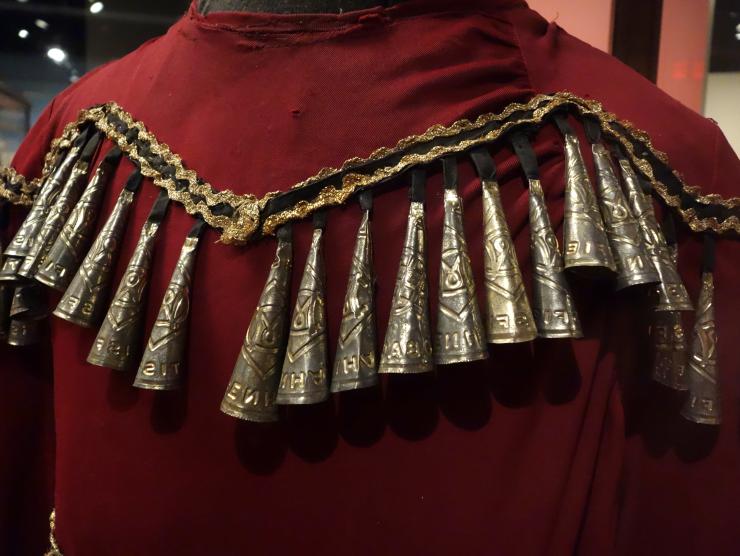 Photo of the back yoke detail of a red jingle dress. Black contrasting trim and gold rick rack in a zigzag pattern across the back shoulders of the dress, and the individual jingle pieces are attached with black fabric. The jingles themselves are each made of the lid of a metal tobacco can, and curled into a conical bell shape. The small end is affixed to the black fabric, and as the dancer moves, the jingle pieces touch to create sound.