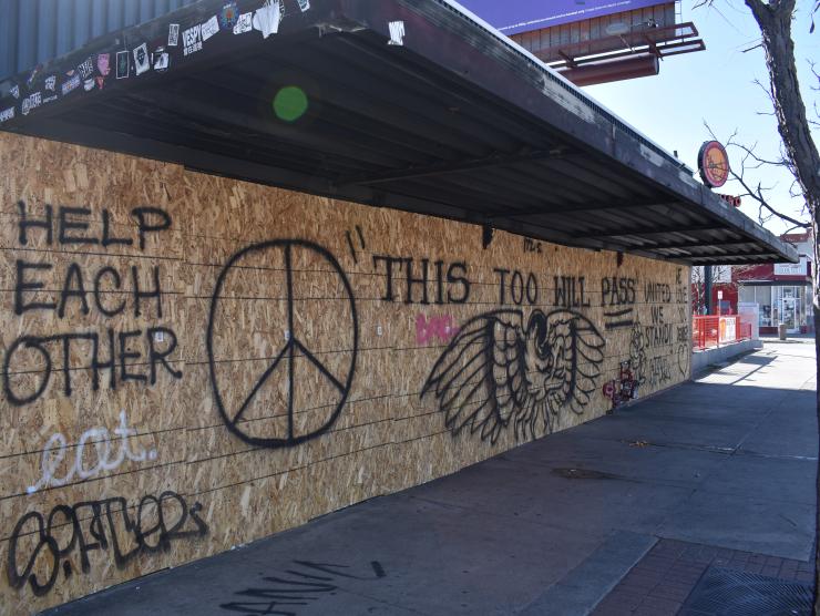 Photo of Think Tank Tattoo, with windows boarded up due to closure during the 2020 pandemic. The main image spray painted on the boards is an eagle and "THIS TOO WILL PASS". Right side detail consists of black outline of a flower, heart and the words, "UNITED WE STAND" and "WE LOVE YOU DENVER!!" 