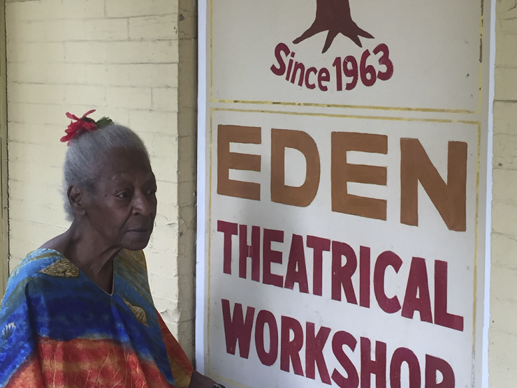 An elderly Black woman, Lucy Walker, is seated in a colorful shawl before a sign which reads Eden Theatrical Workshop (since 1963)
