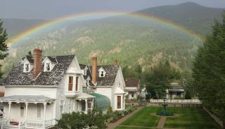 A white house on the left with a well manicured green lawn on the right and a green fountain in the center of the lawn. A rainbow crosses the entire picture above the house intersecting the mountains in the background.