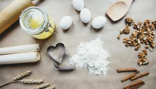 Photo of ingredients used in baking, laid out on a counter. A wooden rolling pin and a roll of parchment, along with a few sheafs of wheat are on the left of the image, while a small mound of flour is next to 2 heart-shaped cookie cutters in the center. Four eggs are nearby, as well as walnuts and stick cinnamon.