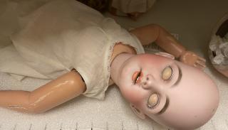 Photo of a doll, laying on a metal shelf lined with a thin sheet of plastic packing. The doll is dressed only in a cotton underdress with no sleeves. The doll has no hair and its eyes are closed, but because of the difference in color of eyelid and face, it appears as though the doll has blank eyes. Its orange-lipped mouth is open, and its arms are askew.