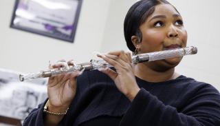 Photo of pop performer Lizzo, holding a flute to her mouth to play. The flute is crystal with silver bands. Lizzo is a Black woman, whose black hair is parted in the center and smoothed, pulled to the back of her head. She is wearing gold earrings and a bracelet, and a navy blue long sleeve sweater. She is standing in a bright white room that has a framed image hanging on the wall behind her. She is looking up toward the ceiling as she plays, her lips pursed as she blows into the instrument.