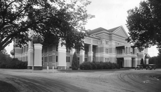 A black and white photo of a large building with geometric features. Its exterior is dominated by pillars and its peaked roof is higher than the trees around it. There is a dirt driveway leading up to the building where a car is parked.