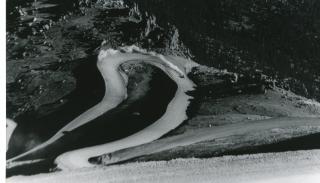 Photo of a portion of the twists and turns of the Pikes Peak International Hill Climb. The view is of a race vehicle having just gone around a hairpin turn, kicking up a trail of dust from the dirt track. Beyond the track lies the rocky hill top of the mountain, the steep terrain evident as the car climbs higher along the course.