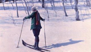 A woman stands on two skis in a snowy landscape. She is wearing a jacket, scarf, and hat, and looks over her shoulder at the camera.