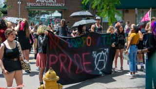 Photo of activists standing in front of a brick building. There is a group of a few dozen people in this image, standing in small groups talking with each other, looking around. A few people in the front carry a sign made of black fabric. The sign says "NO CORPORATE PRIDE" painted in colorful letters, and a large transgender symbol (a pictogram consisting of a circle with an arrow sprouting from the top-right side, a cross at the bottom, and a stroked arrow at the top) painted in white in the lower corner.