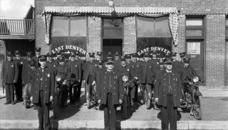 Black and white photo of a group of Denver police officers pose near a building with signage saying East Denver Police Station.