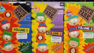 A photograph taken with a cell phone camera, of three VHS tapes in their cardboard covers, lined up on white metal shelving so that the full cover of each tape is visible. The tapes are recordings of the "South Park" animated television series, Volumes 1, 2, and 3. Each cover prominently displays the "TV-MA" warning in the upper right corner, as well as a warning box below the volume number, indicating that these programs are for mature, audiences only and contain adult language and situations.
