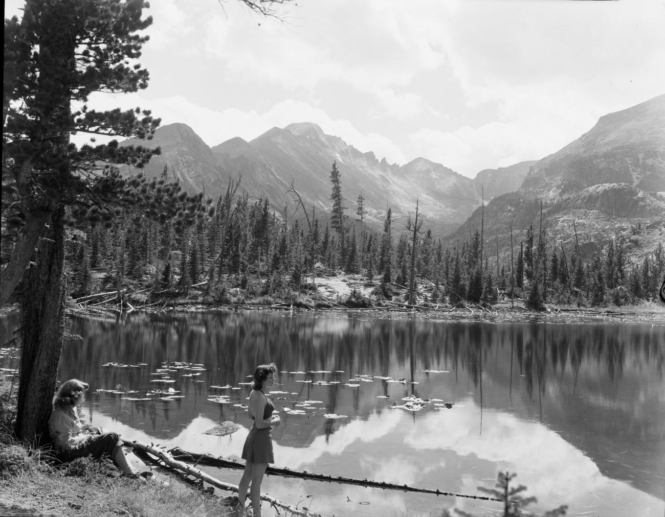 Two women on the bank of Nymph Lake in Rocky Mountain National Park, framed by trees and mountain peaks.