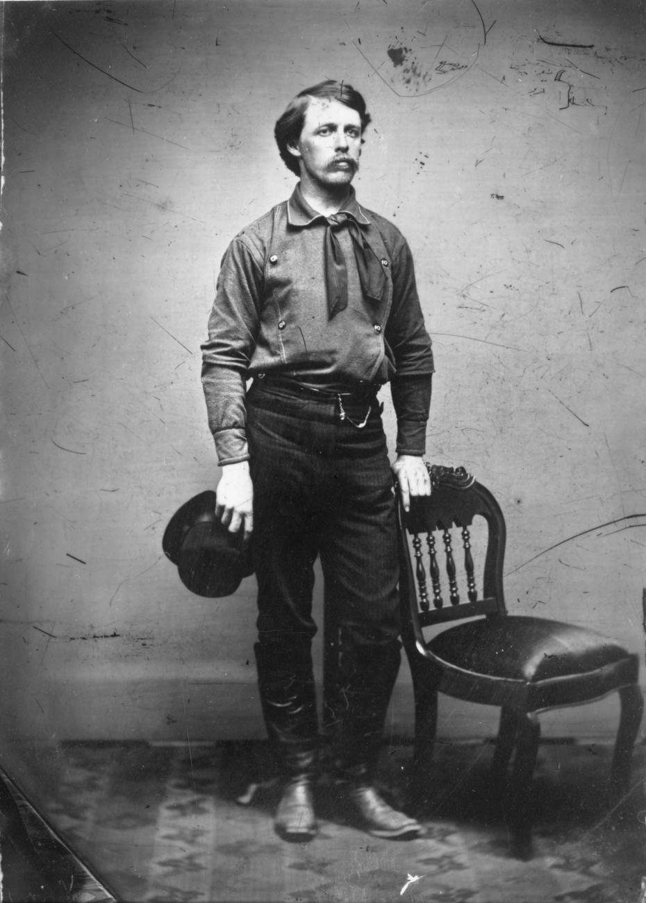William Henry Jackson posing in a studio, his hat in his right hand, his left hand gripping a chair. 